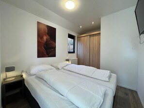 A modern bedroom with a bed, bedside tables, a wall-mounted TV, and a large window with blinds. - Cozy Chalet With Dishwasher, Adjacent to the Hoge Veluwe National Park (Arnhem)