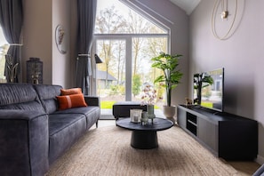 A modern living room with a dark sofa, a round coffee table, and a large window with a view of trees and houses. - Eco Villa in Latour by Scenic Pond (Oirschot)