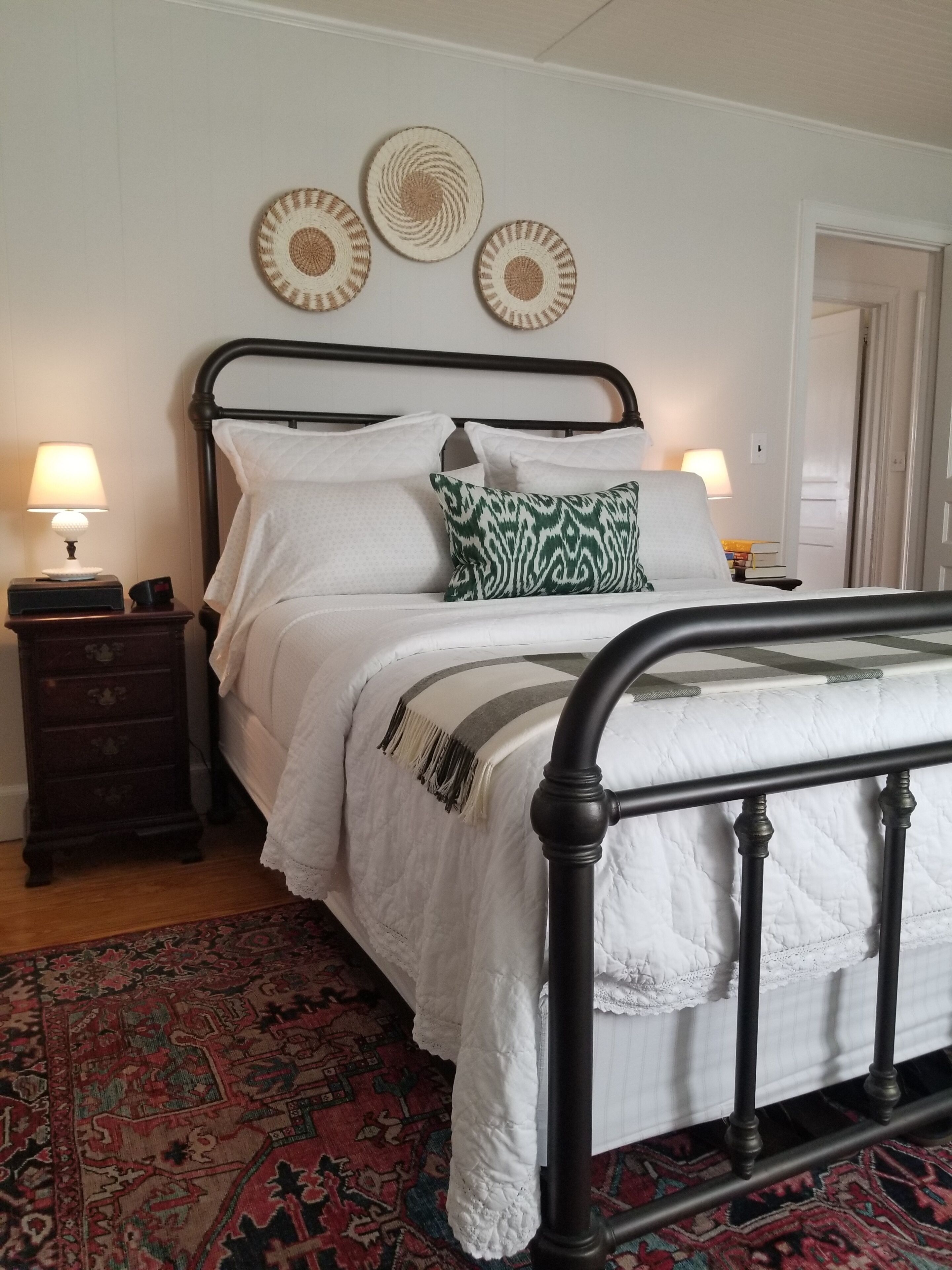 Een netjes opgemaakt bed met witte lakens en een kussen met een groen patroon, een nachtkastje van donker hout met een lamp, en een vloerkleed met een rood patroon.