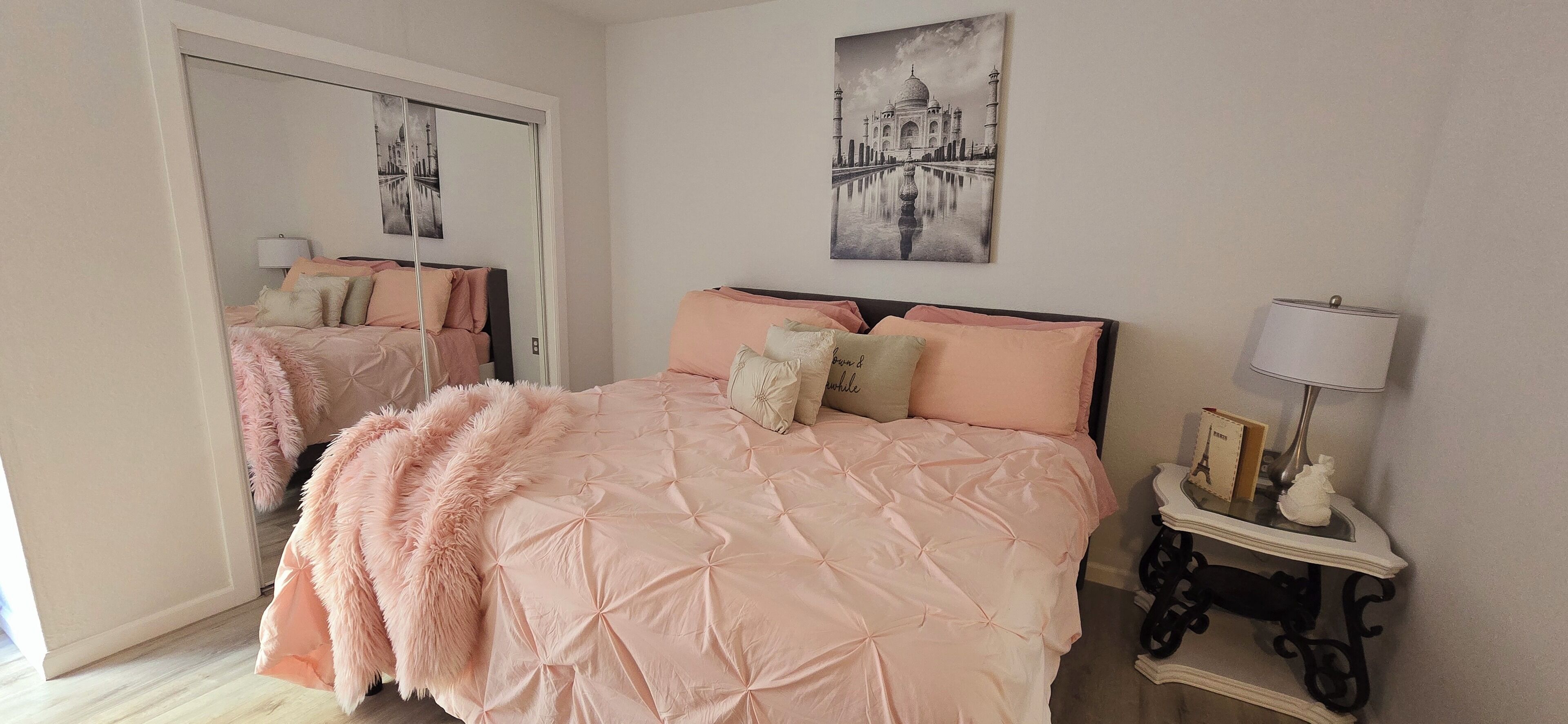 A bedroom with a large bed, a mirror, a picture of the Taj Mahal, and a bedside table with a lamp.