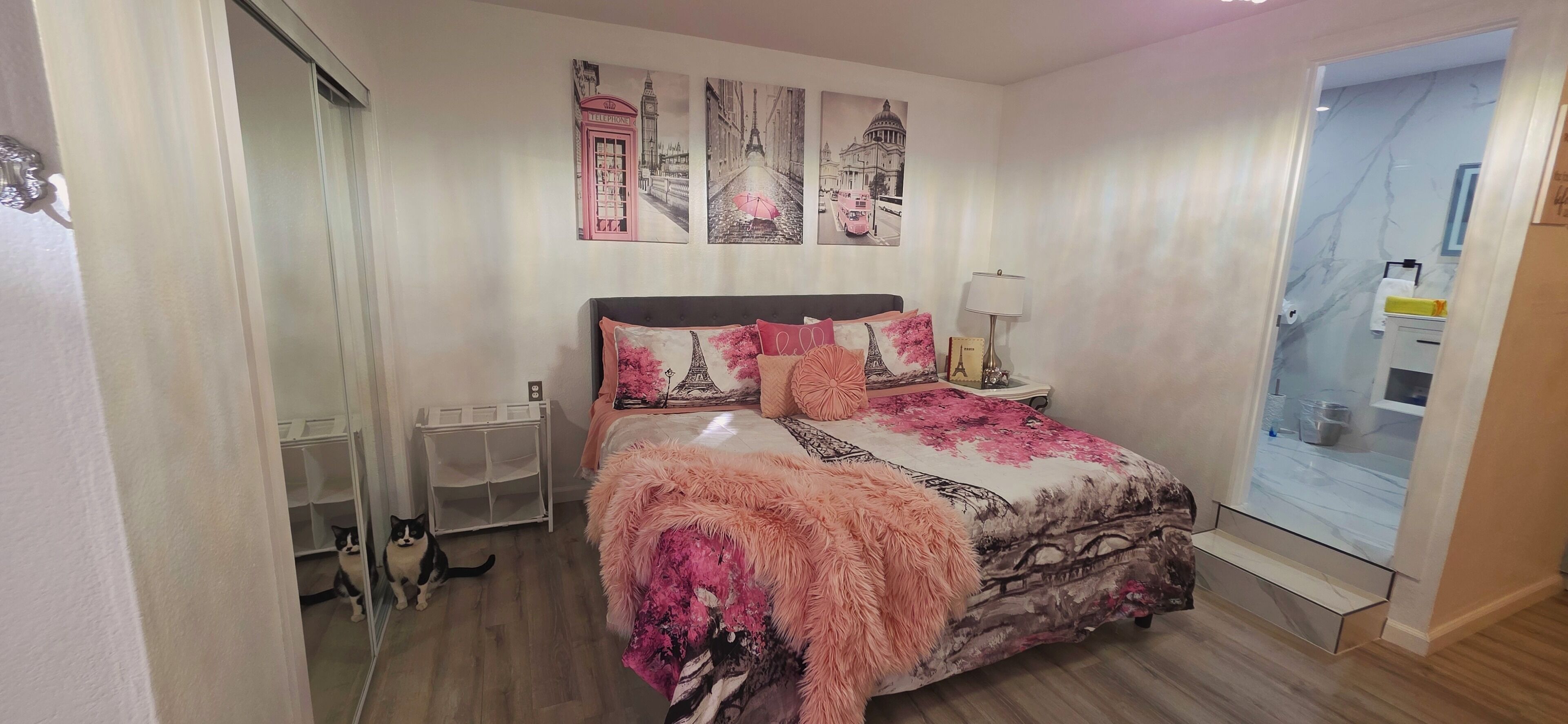 A bedroom with a bed, a nightstand, a lamp, and a mirror. There are wall art pieces above the bed and a door leading to another room.