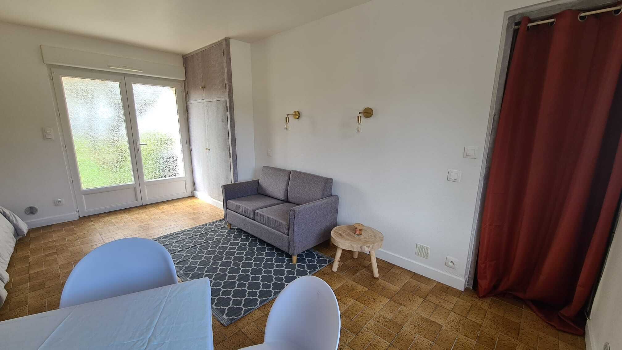 Ein Zimmer mit grauem Sofa, einem runden Holztisch, einem gemusterten Teppich und einer Schiebetür zum Balkon.