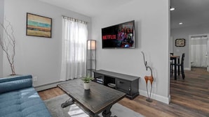 Ein modernes Wohnzimmer mit einer Couch, einem Couchtisch, einem an der Wand befestigten Fernseher und einem Essbereich im Hintergrund.