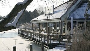 A wooden pier leading to a building with a tiled roof, surrounded by snow-covered ground and trees. - Bungalow on Weerterbergen Waterfront (Weert)