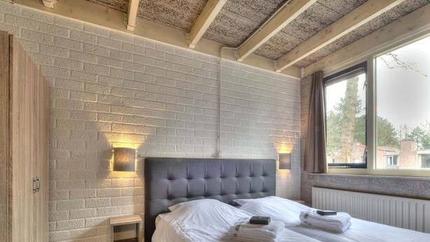 A bedroom with a brick-patterned wall, a tufted headboard, bedside lamps, a wooden wardrobe, and a window with a view of buildings. - Bungalow on Weerterbergen Waterfront (Weert)