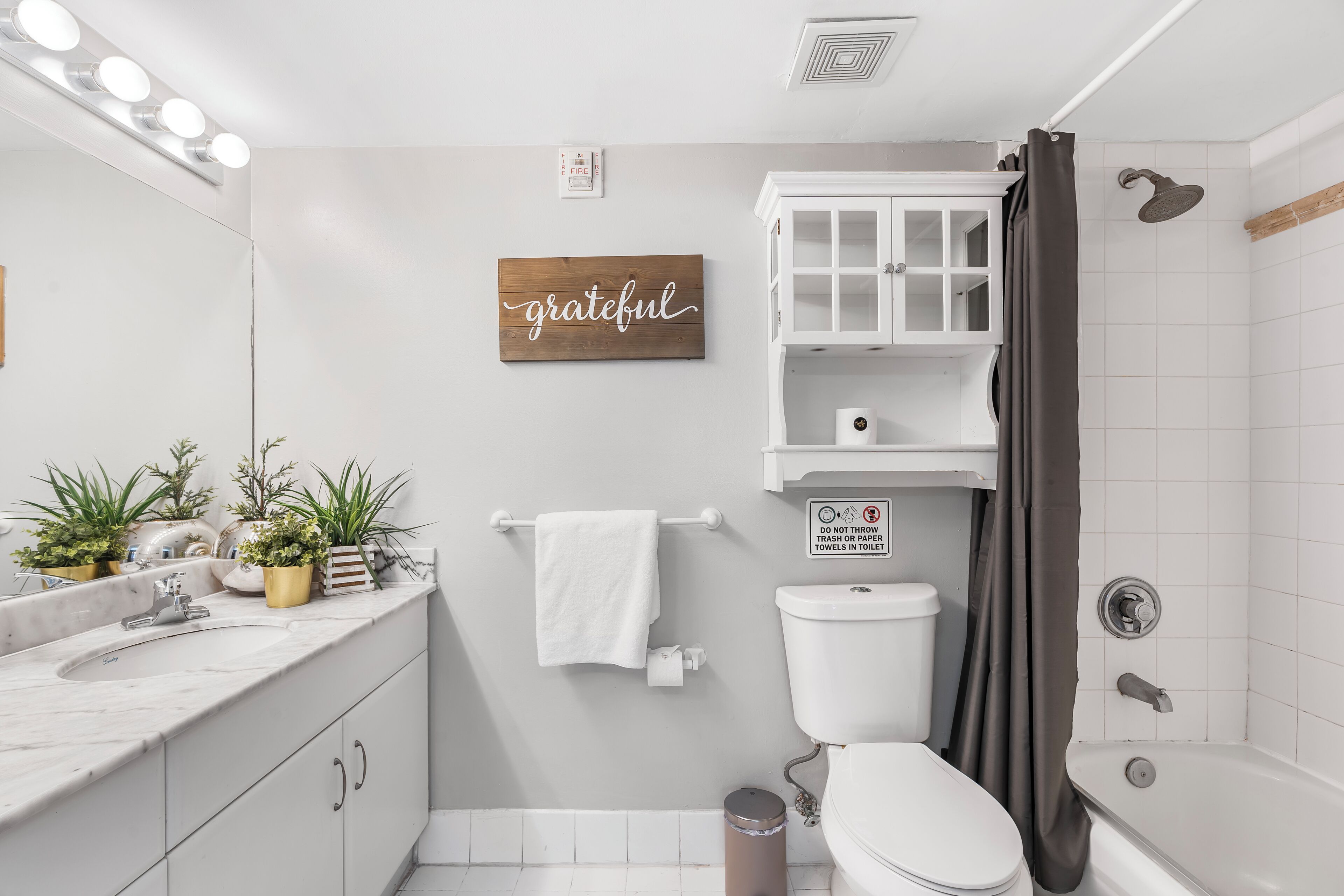 Ein Badezimmer mit weißem Waschbecken, WC und Badewanne. Auf der Ablage stehen Blumentöpfe und ein Schild mit der Aufschrift 'grateful'.