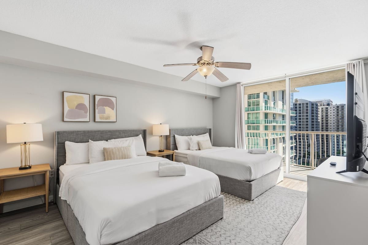 A hotel room with two beds, a ceiling fan, a large window with a view of high-rise buildings, and a desk with a television.