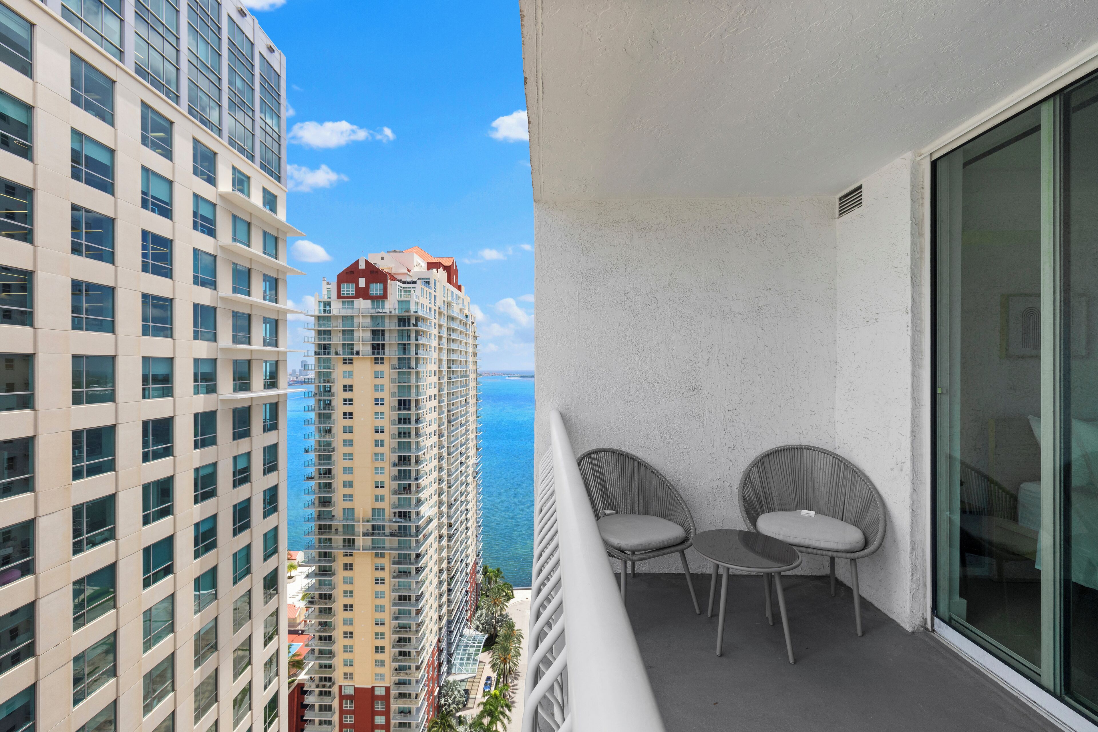 A balcony with two chairs and a table, overlooking a high-rise building and the ocean.