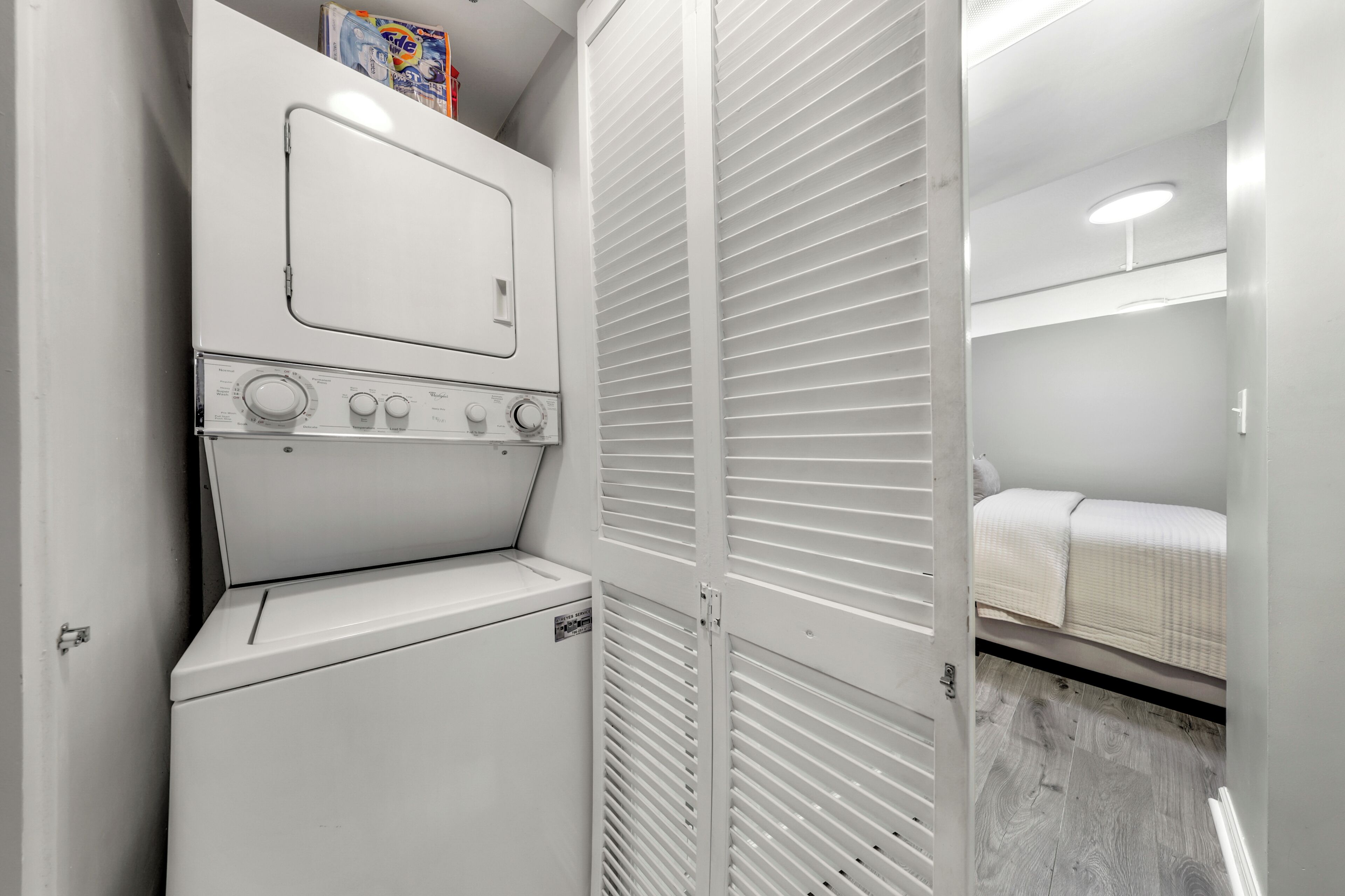 A compact laundry room with a washer and dryer, a white closet with louvered doors, and a bunk bed with a white mattress.