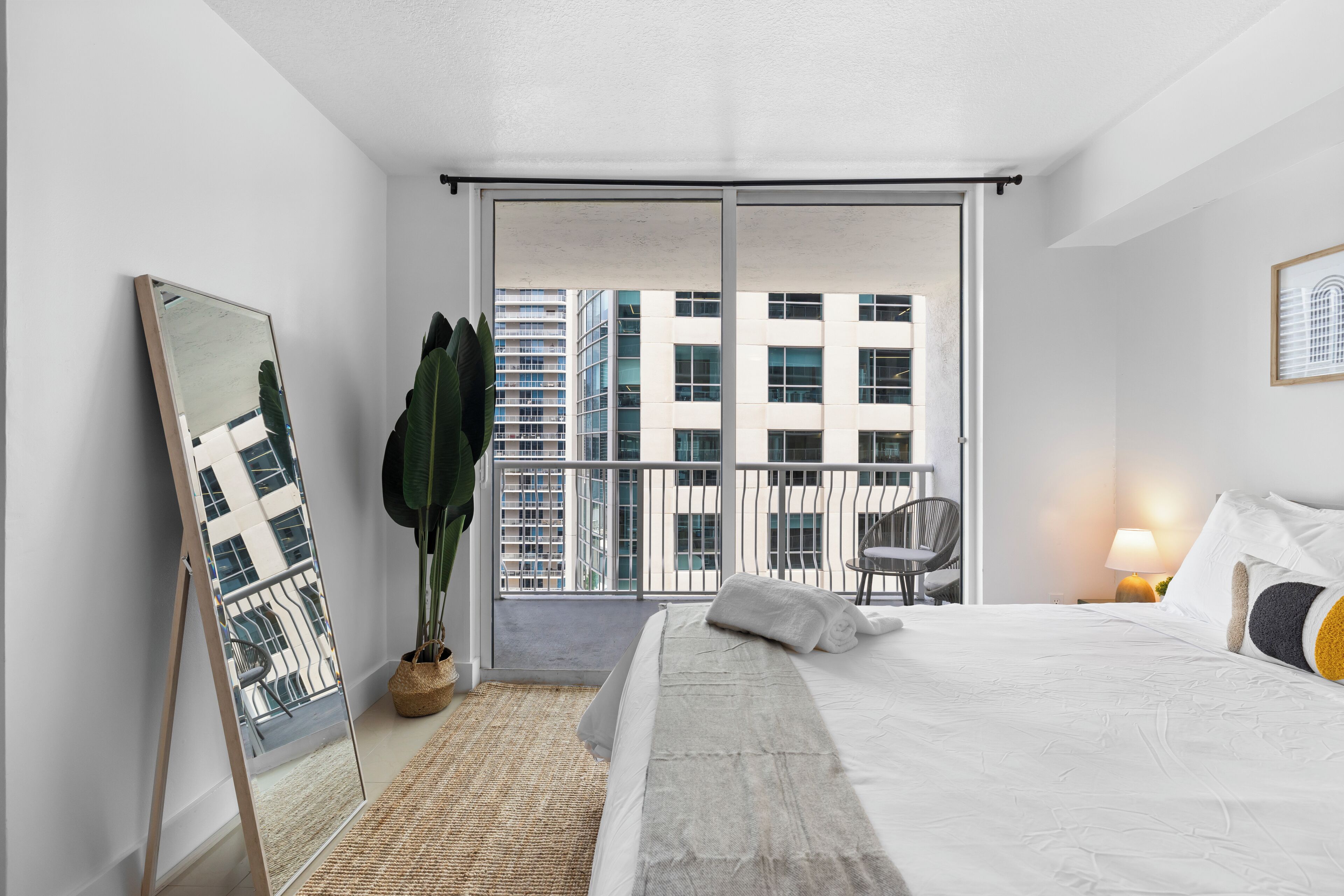 Une chambre d’hôtel moderne avec un grand lit, un miroir, une plante en pot et une vue sur le paysage urbain, visible à travers une porte coulissante.