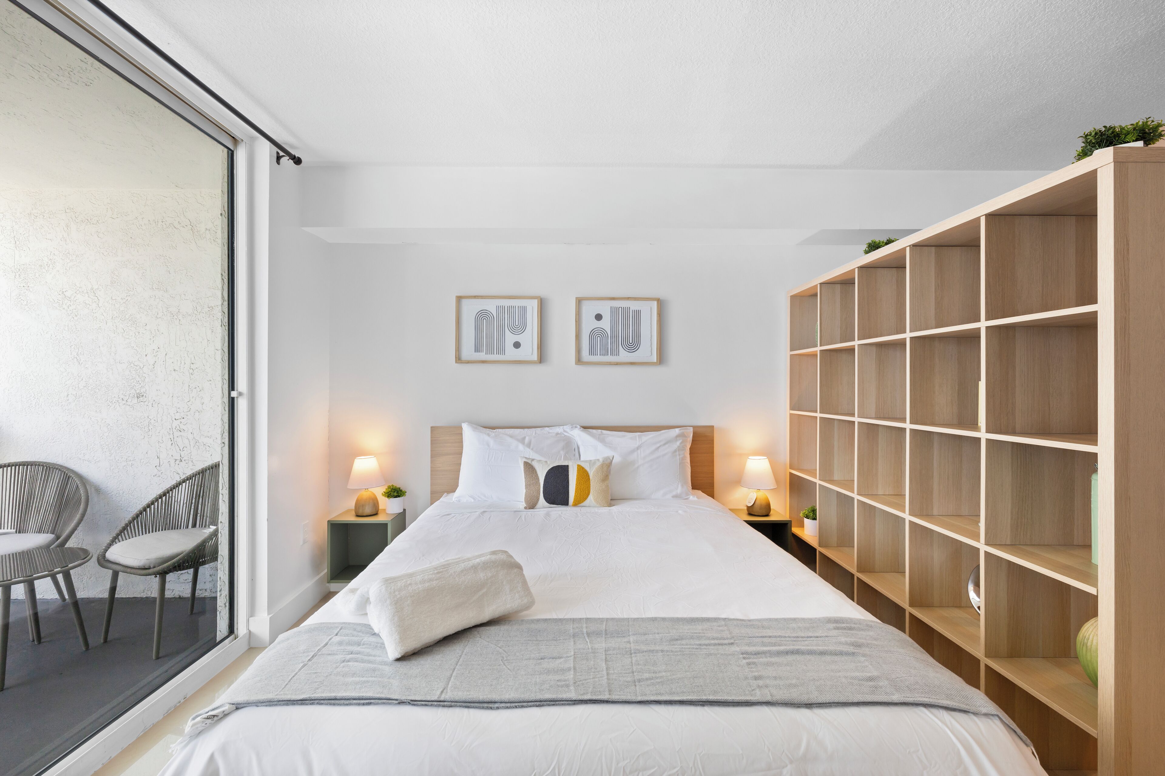 A modern bedroom with a large bed, two bedside tables with lamps, a built-in wooden shelving unit, and a sliding glass door leading to a balcony with wicker chairs.