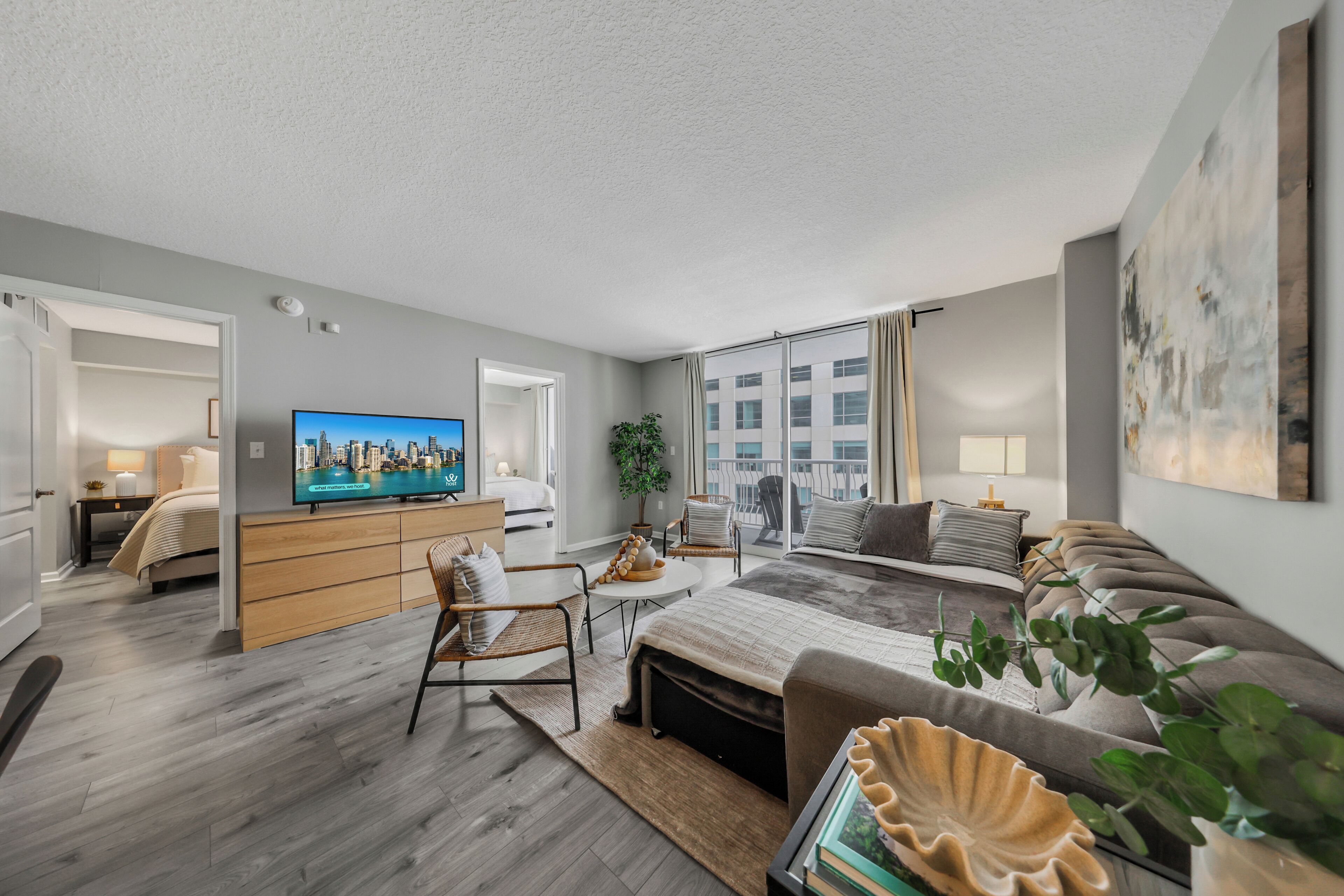 A modern hotel room with a large bed, a TV, a dining area, and a view of the city.