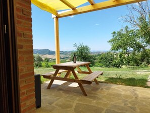 Een houten picknicktafel onder een geel dak, op een stenen terras met uitzicht op glooiende heuvels en wijngaarden.