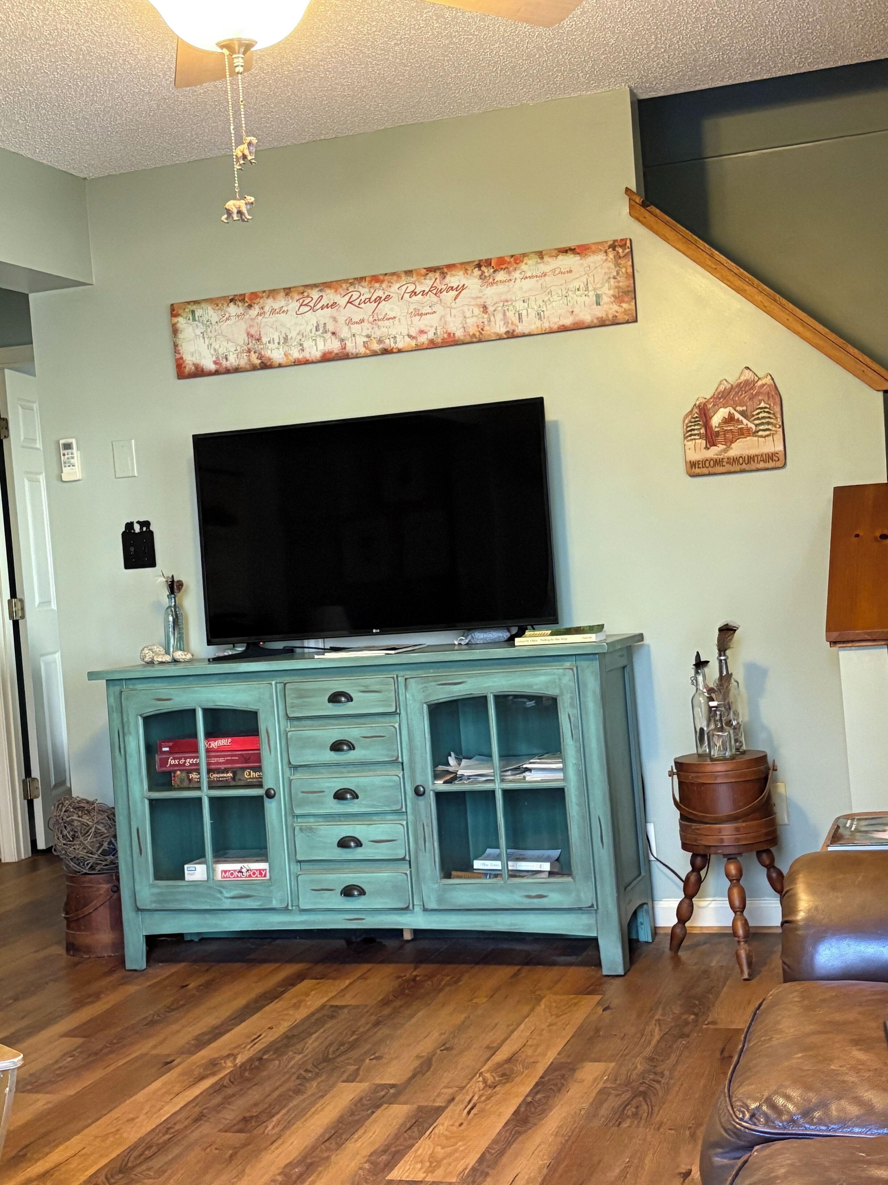 Een woonkamer met een blauwgroen kastje, een flatscreen televisie, houten vloeren en een trap.
