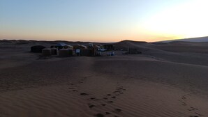 Paisaje desértico con dunas de arena, un pequeño asentamiento con edificios y un vehículo, e huellas en la arena.