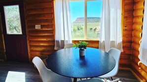 Une pièce de cabane en bois avec une table ronde, des chaises blanches, une plante en pot et une fenêtre avec des rideaux.