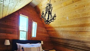 Une chambre confortable avec un plafond en pente en bois, un lit recouvert de draps blancs, deux fenêtres et un luminaire suspendu.
