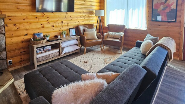 L’intérieur d’une cabane en rondins, avec un grand canapé, deux fauteuils, une tête de cerf accrochée au mur, un téléviseur à écran plat et une cheminée en pierre.