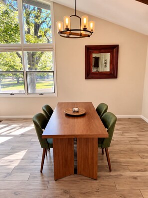 A dining area with a wooden table, green upholstered chairs, a chandelier, and a large window with a view of trees.