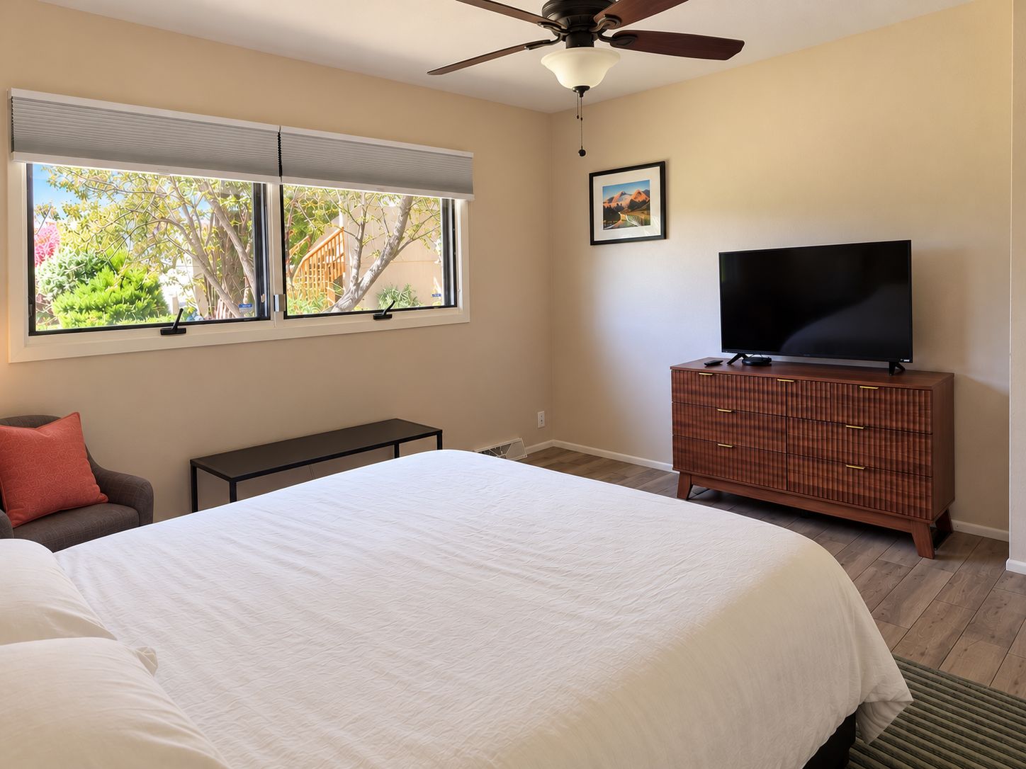 A bedroom with a bed, a television on a wooden cabinet, a ceiling fan, and a window with blinds.