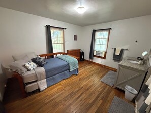 Ein Schlafzimmer mit einem Bett, einer Kommode, einem Schminktisch mit Spiegel und zwei mit Vorhängen versehenen Fenstern.