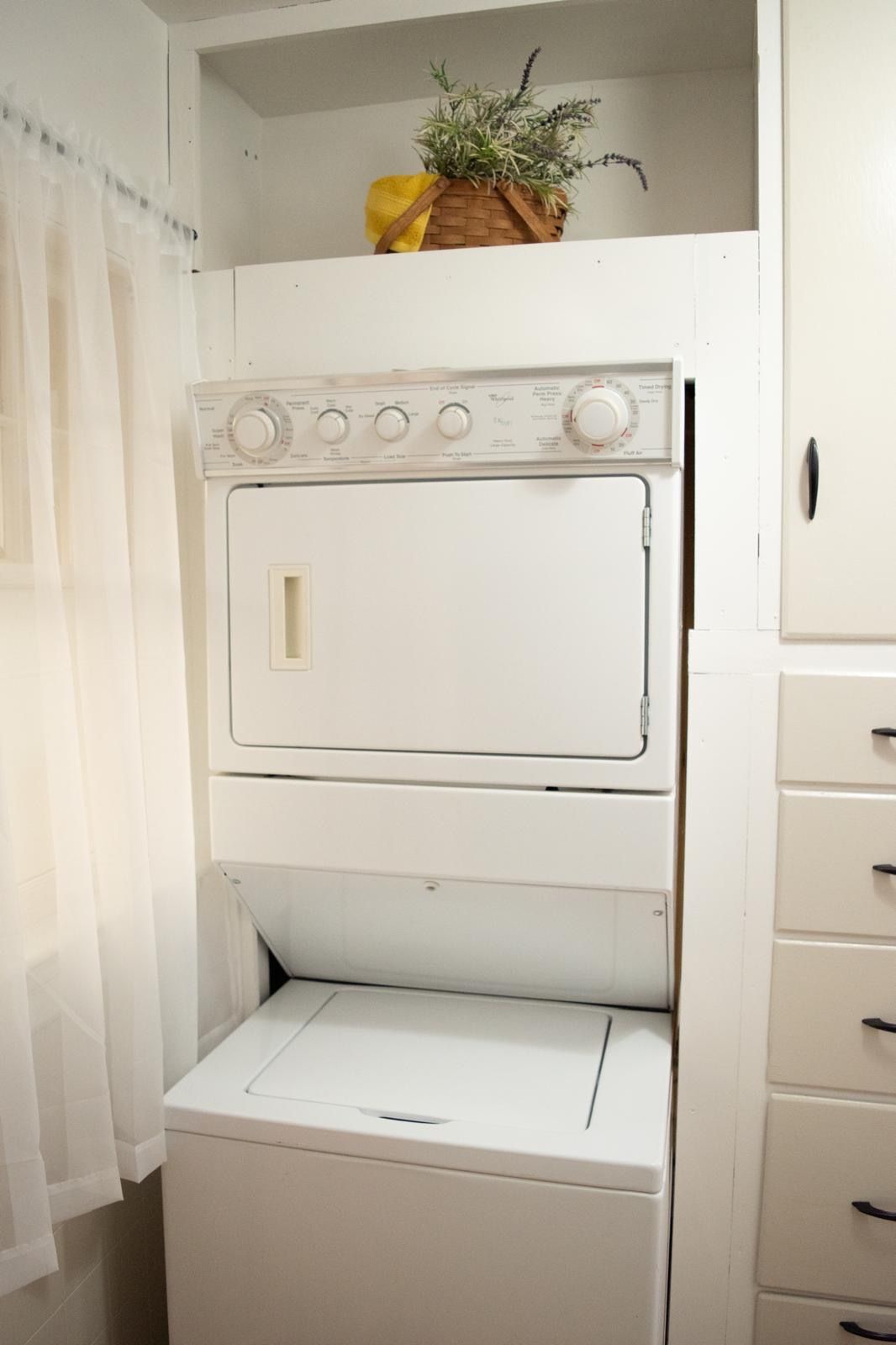 Un lavadero blanco con lavadora y secadora, una cesta de madera con plantas, y un armario blanco con cajones.