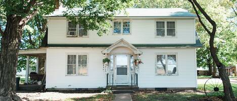 A two-story white house with a front porch, a small front yard, and a gravel driveway.