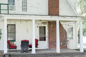 Een wit huis met een veranda, twee rieten stoelen met rode kussens en een bakstenen schoorsteen.