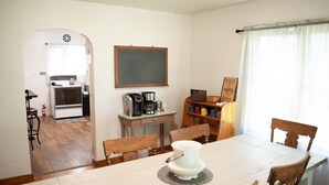 A dining area with a white table, wooden chairs, a coffee station, and a view of a kitchen with an oven and a refrigerator.