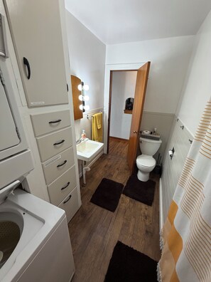 Een kleine badkamer met een wit wastafelmeubel, toilet en een spiegel met verlichting. De vloer is van hout en er is een handdoekhouder.