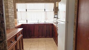 A kitchen with wooden cabinets, a white refrigerator, and a window with lace curtains. - Sandcastle: 6x2 home with stunning views of both the ocean and the sand dunes  (Lancelin)