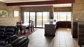 A spacious living area with a dining table, chairs, a black leather sofa, a kitchen with a brick backsplash, and a sliding glass door leading to a patio. - Sandcastle: 6x2 home with stunning views of both the ocean and the sand dunes  (Lancelin)