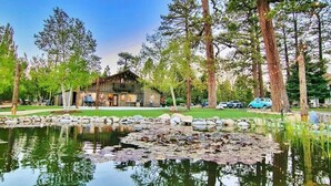 Un chalet rustique avec un étang devant, entouré de grands arbres et sous un ciel dégagé.