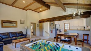 Une pièce comprenant une table de baby-foot, un canapé, une table basse et un coin cuisine avec des poutres en bois et un lustre.