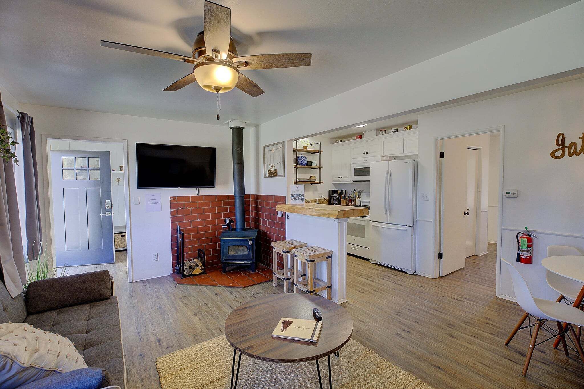 A living room with a sofa, a round table with a book, a ceiling fan, a fireplace, and a kitchen area with a refrigerator and a microwave.