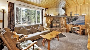 A cozy living room with a stone fireplace, wooden furniture, and a large window offering a view of trees. - Reality Escape #2466 (Big Bear City)