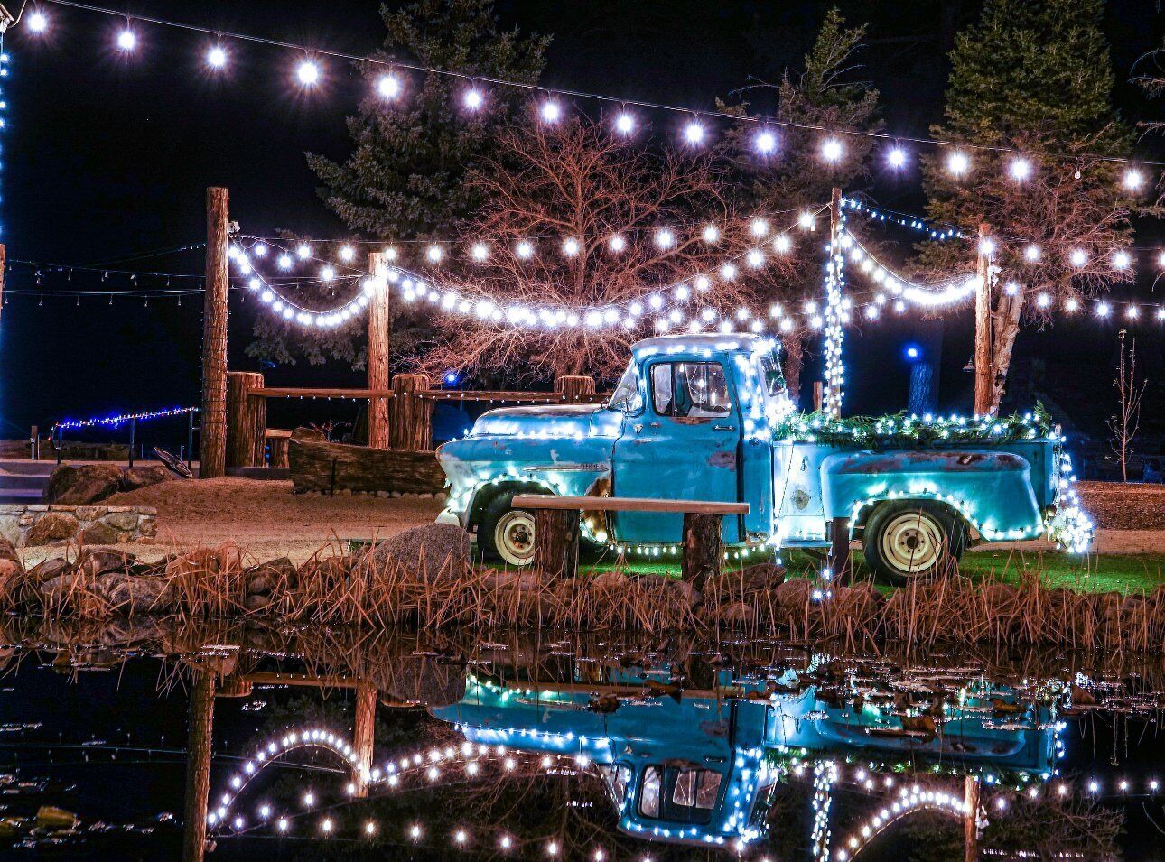 Una camioneta azul antigua decorada con luces, estacionada junto a un cuerpo de agua con reflejos.