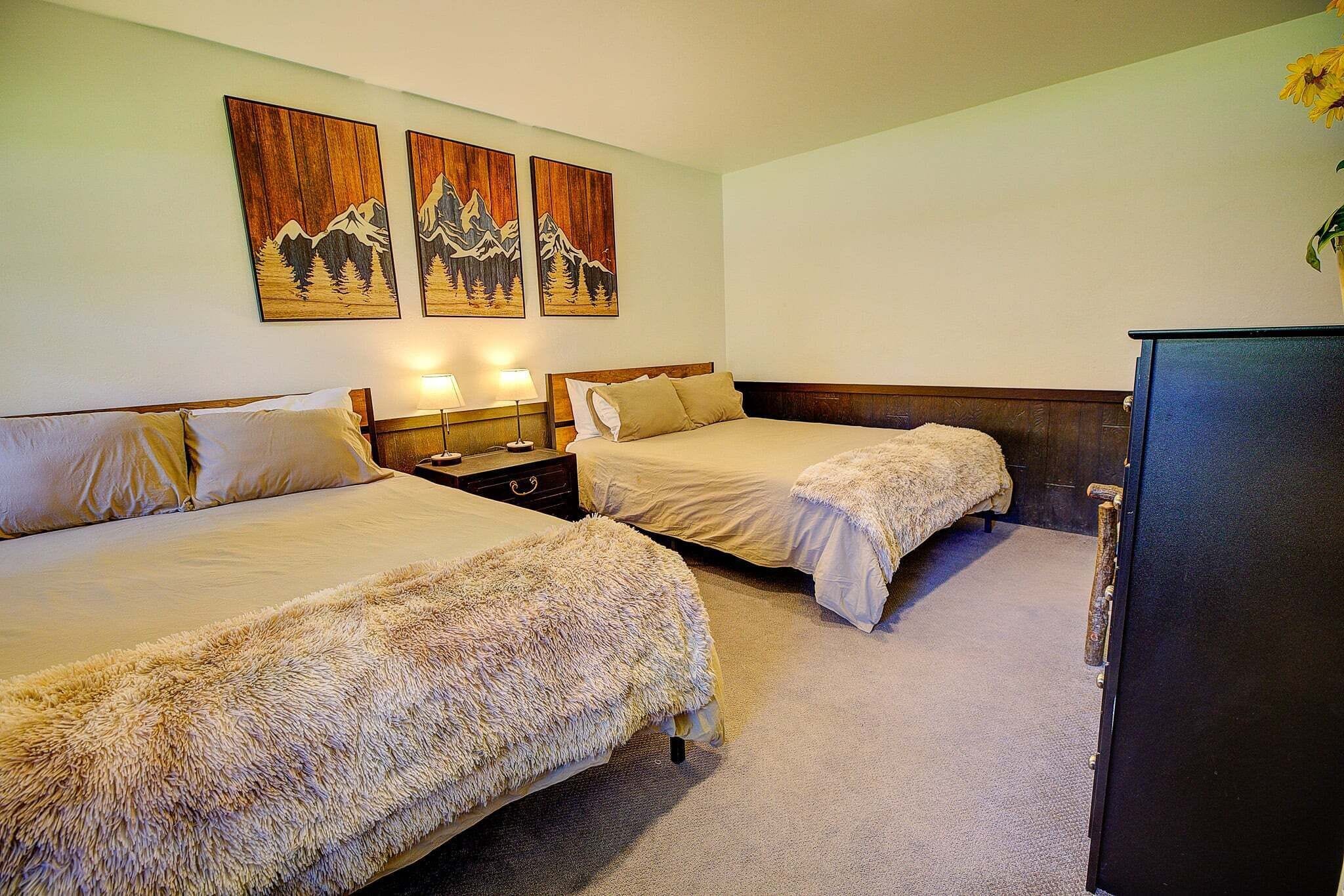 A hotel room with two beds, a nightstand, a dresser, and wall art depicting mountains.