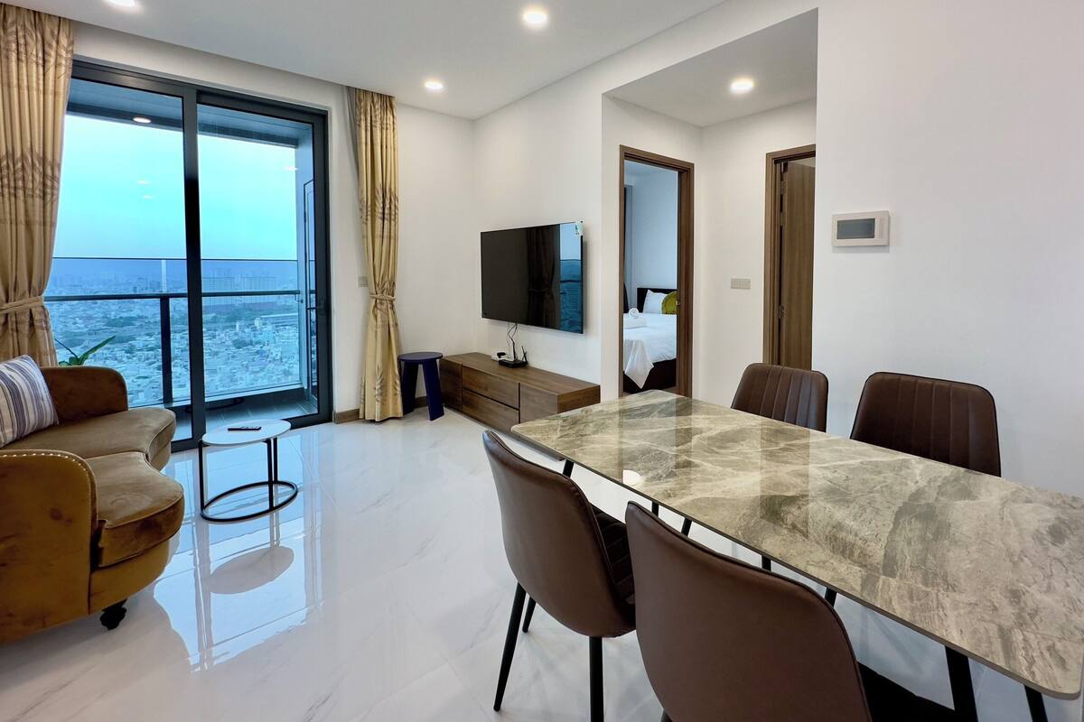 Une chambre d’hôtel moderne dotée d’un grand balcon, d’une table à manger avec des chaises, d’un téléviseur fixé au mur et d’un lit visible à travers une porte ouverte.