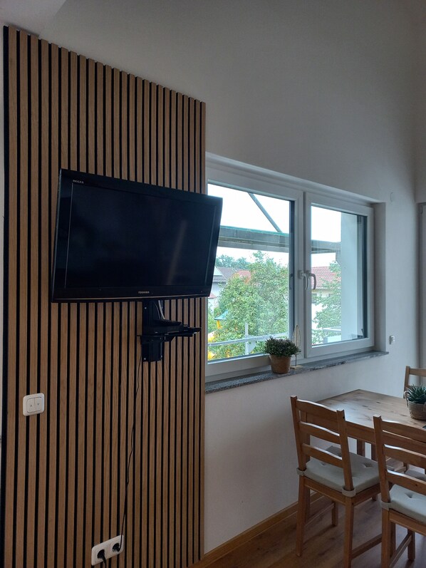 A room with a flat-screen TV mounted on a wooden slat wall, a dining table with chairs, and a window with a view of greenery and buildings outside.