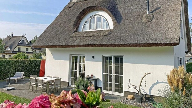 Une maison avec un toit de chaume, des murs extérieurs blancs et un petit jardin agrémenté d’une table et de chaises.