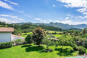 A rural landscape with a white building, green fields, and mountains in the background.
