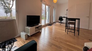A spacious living area with a flat-screen TV on a white stand, a dining table with chairs, and a pendant light.