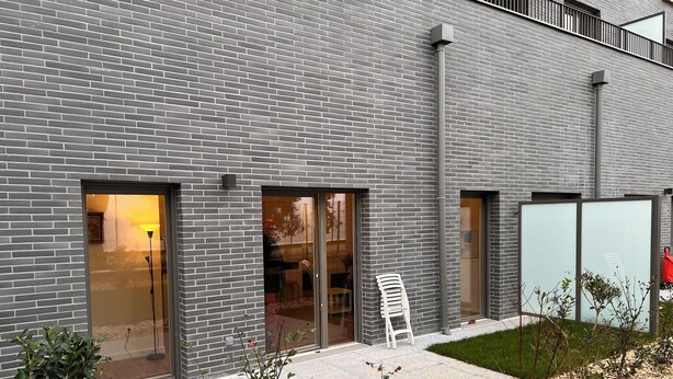 A modern building with grey brick walls, large glass doors, and a small outdoor seating area with a white chair.