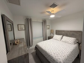 Une chambre à coucher comprenant un lit, une table de chevet, un miroir, un ventilateur de plafond et une fenêtre avec des rideaux.