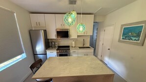 Une cuisine moderne avec des meubles blancs, un îlot central, deux tabourets de bar, un réfrigérateur en acier inoxydable, une cuisinière et des luminaires suspendus.