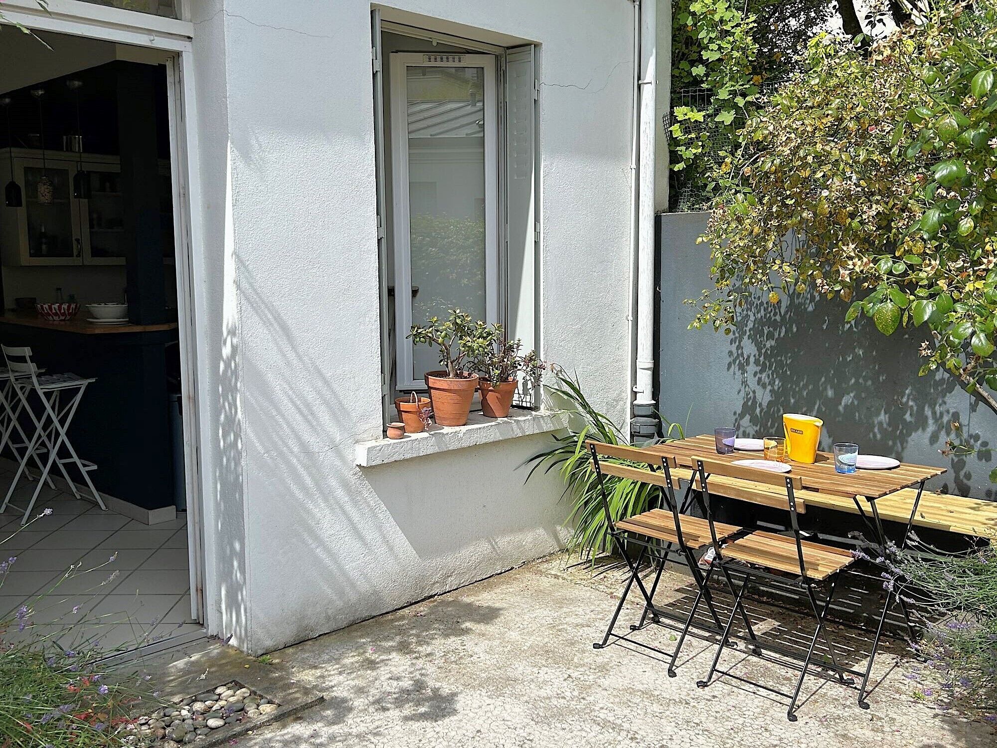 Une terrasse avec une table et des chaises en bois, une fenêtre ornée de plantes en pot et une vue sur un bâtiment dont on aperçoit la cuisine à travers la fenêtre.