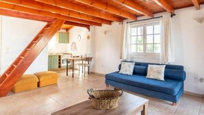 Ein gemütliches Wohnzimmer mit einem blauen Sofa, Holzbalkendecke, einem Essbereich und einer Küche im Hintergrund.