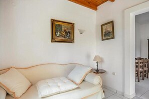 Quarto com sofá branco, duas imagens emolduradas na parede, uma mesinha com abajur e teto de madeira.