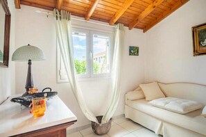Quarto com teto de madeira, sofá branco, mesa com abajur e janela com cortinas.
